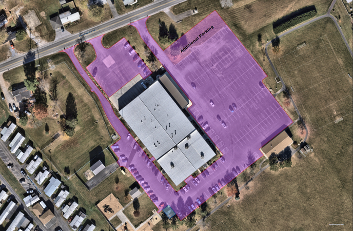 Aerial view of a large institutional building and surrounding grounds, with all existing and proposed paved areas—including driveways, parking lots, and adjacent additional parking zones—highlighted in semi-transparent purple overlay, showing access points on the north, east, south, and west sides.