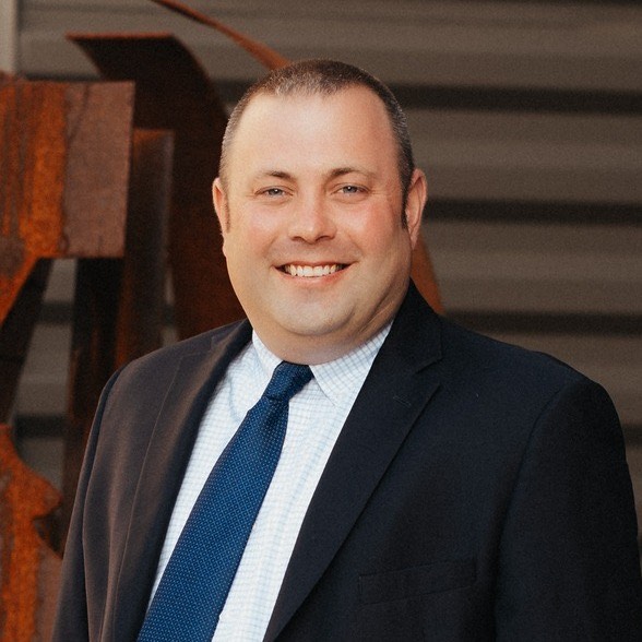 Headshot of Justin Eberly wearing a dark suit jacket, white checkered dress shirt, and blue tie, smiling while standing in front of an industrial-style background with a rust-colored metal structure.